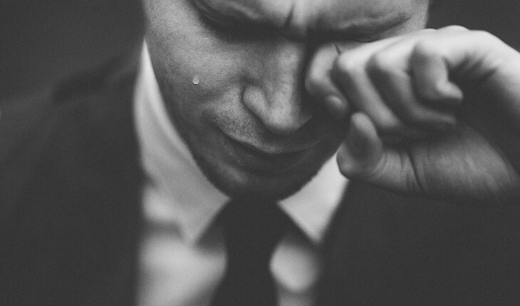 Close-up of a man in a suit looking down while wiping a tear from his eye as he cries due to a painful popping jaw