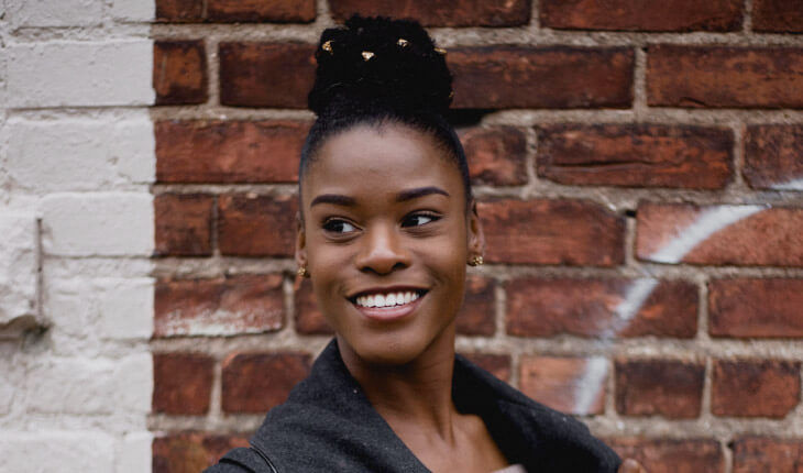 A woman smiles with professionally whitened teeth against a red brick wall with her dark hair in a bun atop her head