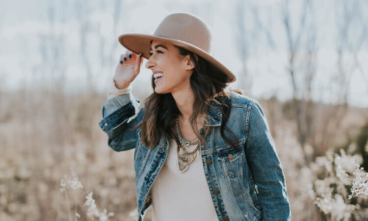 Brunette woman wears a denim jacket, white blouse, necklace, and beige hat as she smiles in a field of tan shrubbery