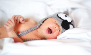 Young baby wearing a knitted raccoon hat sleeps on white sheets, with their first tooth erupting in a few months