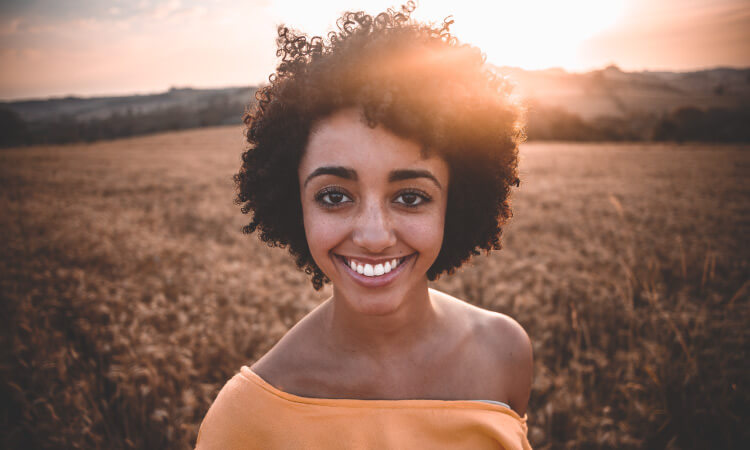 Young woman with dark curly hair wearing a yellow blouse smiles while standing in a golden field as the sun sets
