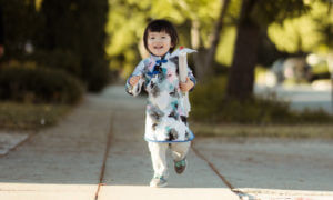 Little girl wearing a white and blue kimono shirt runs along the sidewalk smiling and holding an umbrella