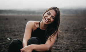 Brunette woman with white straight teeth smiles while wearing a black tank top sitting in a dirt field