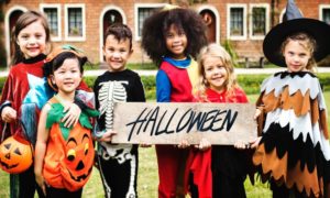 6 smiling children dressed up for Halloween hold a wooden sign that says Halloween