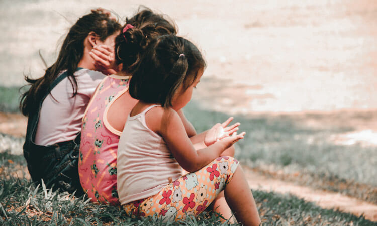 3 dark-haired female little kids wearing floral clothes sit on a creek bank