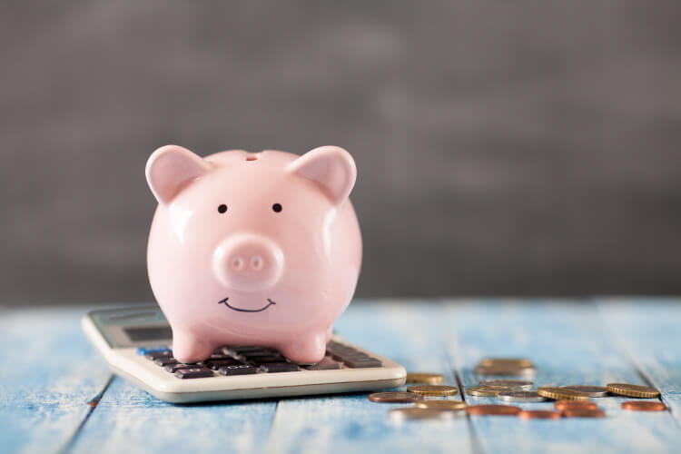 A pink piggy bank used for saving money sits atop a calculator next to a pile of coins