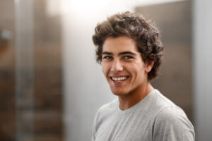 Brunette man in a gray shirt smiles because he utilizes the in-house dental savers plan at Queensboro Plaza Dental Care