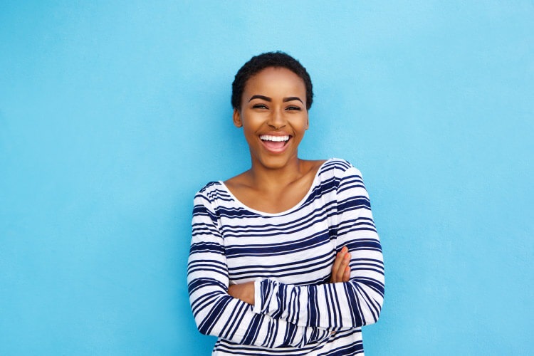 Dark-Haired woman with resin composite fillings smiles while wearing a navy striped shirt against a sky blue wall