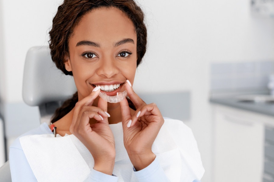 Pretty smiling black girl in the dental chair holding a clear aligner in front of her face.